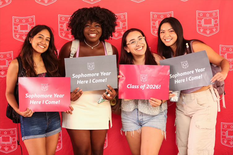 Four students holding first day signs while standing in front of a repeating shield background